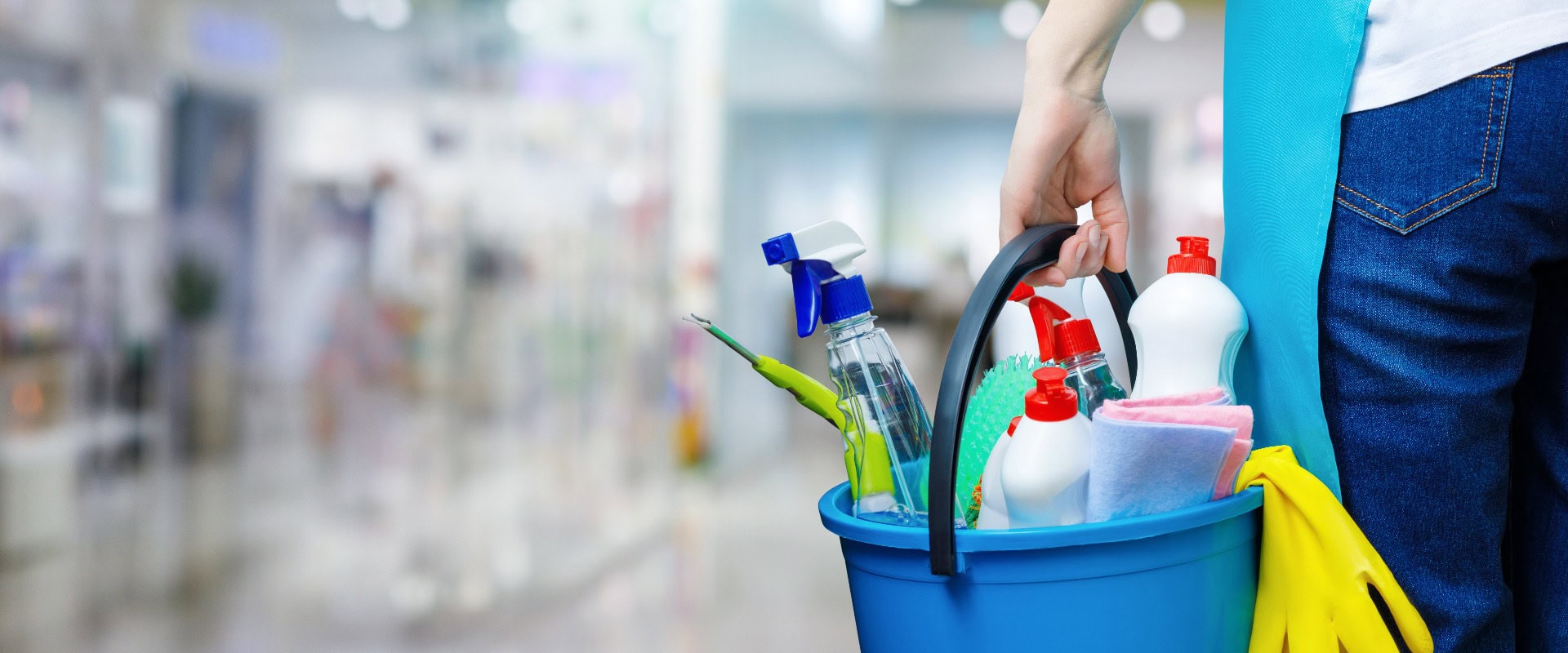 Eine Putzfrau steht in einem Gebäude und hält einen Eimer, der mit Chemikalien und Aufräumutensilien gefüllt ist.